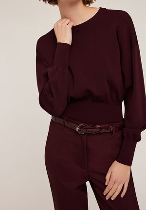 Femme portant un pull en maille bordeaux foncé à manches longues et un pantalon assorti, accessoirisé d'une ceinture tressée sombre et brillante.
