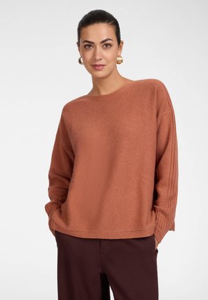 Mujer con cabello oscuro, con un suéter marrón de manga larga y pantalones oscuros, de pie frente a un fondo blanco y liso.