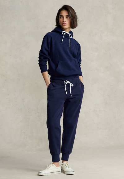 Νεαρό άτομο στέκεται με τα χέρια στις τσέπες, φορώντας σκούρο μπλε φούτερ με κουκούλα και φόρμες με λευκά κορδόνια και λευκά αθλητικά παπούτσια, ουδέτερο φόντο.