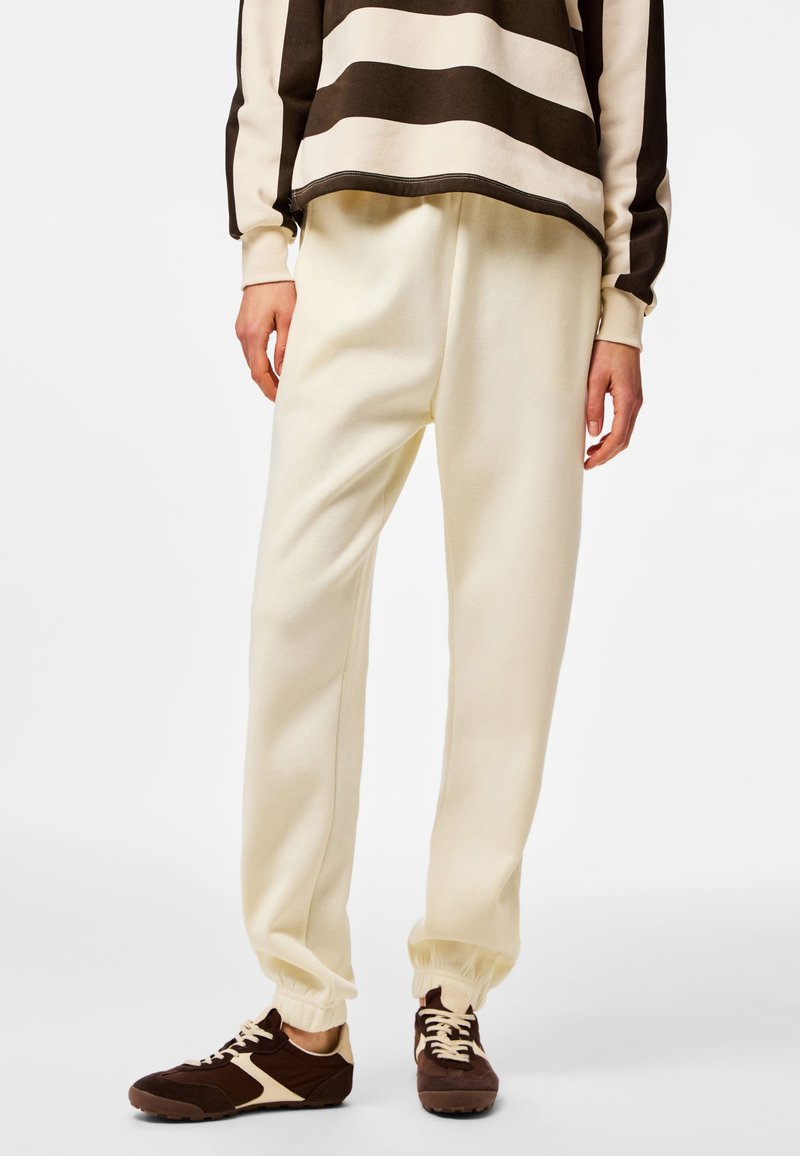 Oseba oblečena v kremasto-barvne tekaške hlače, rjave in kremaste superge ter rjavo-kremasto črtasto majico z dolgimi rokavi, stoji pred belim ozadjem.