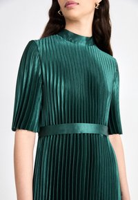 Smaragdgroene geplooide jurk met een hoge kraag en korte mouwen. Heeft een satijnen band in de taille, die een gladde, glanzende textuur toont.