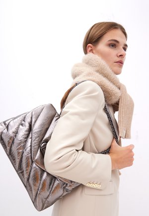 Une femme aux longs cheveux bruns porte un blazer crème et une écharpe beige duveteuse, tenant un sac à motif métallique devant un fond blanc.