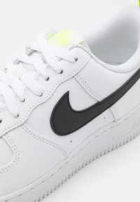 Vit Nike Air Force 1-sneaker med en svart Swoosh-logotyp, strukturerad läderöverdel, platta vita snören och en gr ön detalj på tungan.