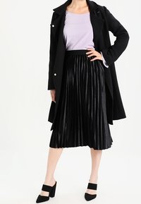 Schwarzer, plissierter Rock kombiniert mit einer helllila Bluse und einem schwarzen Mantel, der mit Knöpfen und einem drapierten Design ausgestattet ist. Schwarze High-Heels.