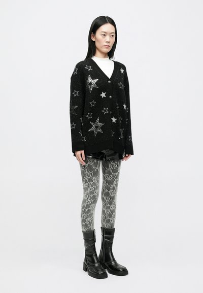 Cardigan negru cu modele de stele argintii, purtat peste un top alb și pantaloni scurți negri strălucitori. Colanți gri cu imprimeu de șarpe și cizme negre până la genunchi.