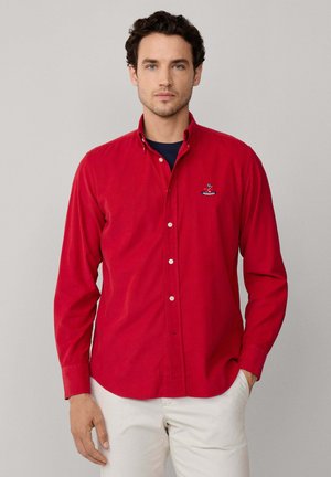 Camicia rossa a maniche lunghe con bottoni in cotone. Presenta un colletto, un logo ricamato e bottoni bianchi. Indossata sopra una T-shirt blu navy.