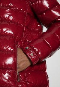 Red puffer jacket with a glossy finish, featuring quilted sections, a zippered pocket, and snap-button cuffs. Smooth texture and fitted design.