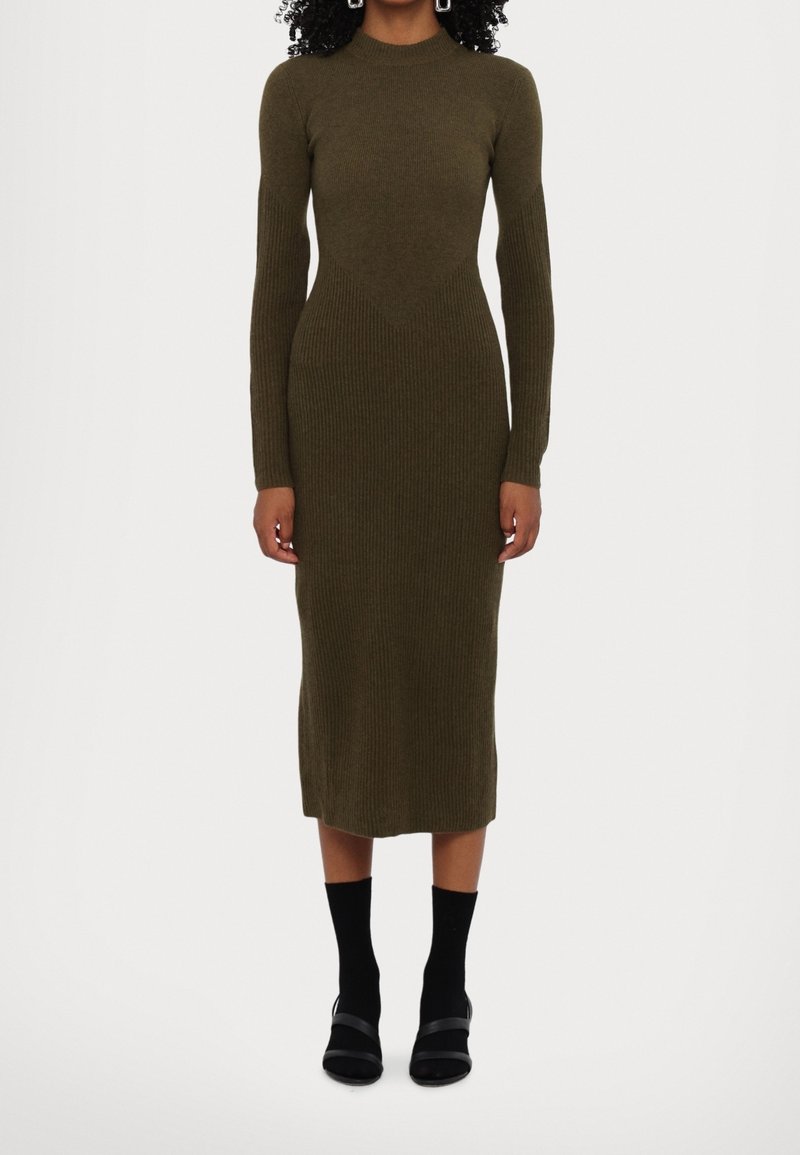 Femme portant une robe midi moulante en tricot côtelé vert olive à manches longues, associée à des chaussettes noires et des sandales noires à bout ouvert.