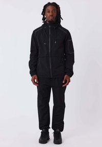 Giacca nera impermeabile con colletto alto, cordino regolabile e chiusura con zip; abbinata a pantaloni neri coordinati e calzature scure.