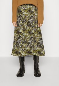 Jupe midi fleurie avec des motifs verts, bruns et crème ; tissu doux qui tombe délicatement. Associée à des bottes noires montantes et un pull brun côtelé.