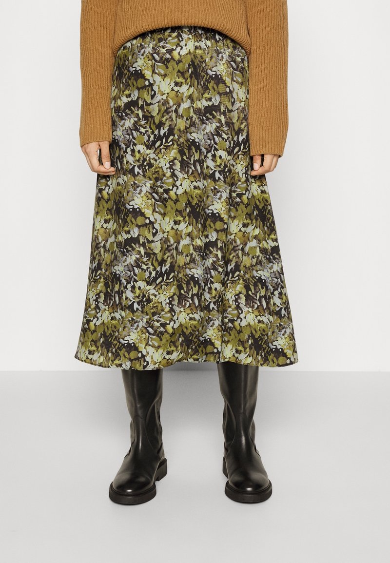 Jupe midi fleurie avec des motifs verts, bruns et crème ; tissu doux qui tombe délicatement. Associée à des bottes noires montantes et un pull brun côtelé.