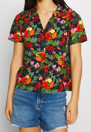 Frau trägt ein marineblaues Kurzarmshirt mit einem leuchtend roten, orangefarbenen und pinkfarbenen Blumenmuster und hellblaue Jeansshorts mit ausgefransten Kanten.