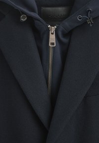 Marinblå jacka med en strukturerad mönster, med dragkedja och huva. Silverfärgad dragkedja och "Polo Club" etikett syns.