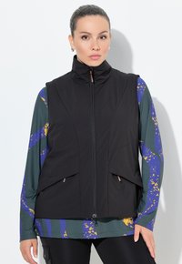 Schwarze ärmellose Weste mit hohem Kragen, ausgestattet mit zwei Fronttaschen und einem durchgehenden Reißverschluss. Darunter ein gemustertes Langarmhemd in Grün und Blau.