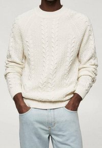 Weißer Strickpullover mit Zopfmuster, dicken gerippten Bündchen und Saum. Kombiniert mit hellblauer Denim-Jeans für einen lässigen Look.