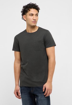 Mężczyzna o kręconych włosach, ubrany w ciemnoszarą koszulkę z kieszonką i niebieskie dżinsy, patrzący w lewo na jednolitym tle.