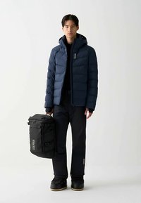 Temno modra puhovka, črne hlače in robustni škornji; stoječi moški drži črno torbo z logotipom. Gladek tekstura, topla zasnova, priložnostna prileganje.