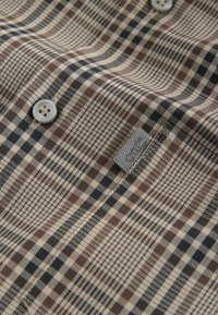 Tissu à carreaux marron, noir et beige avec deux boutons et une petite étiquette en tissu grise cousue près d’une couture.