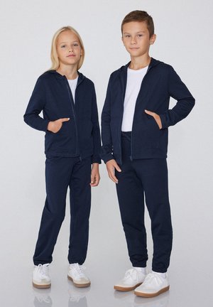 Zwei Kinder tragen marineblaue Kapuzenjacken mit Reißverschluss, passende Hosen, weiße Hemden und weiße Turnschuhe und stehen vor einem einfarbigen Hintergrund.