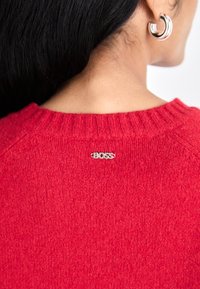 Pull en tricot rouge avec un col côtelé, arborant une étiquette de logo argentée "BOSS" à l'arrière. La texture semble douce et chaude.