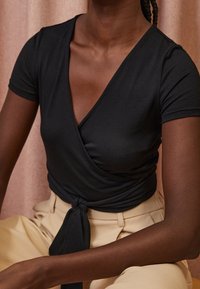Personne à la peau foncée portant un haut cache-cœur noir noué à la taille et un pantalon beige clair, assise devant un fond rose doux.