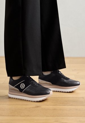 Sorte platform-sneakers med beige og hvide såler, båret med sorte bukser med vide ben på trægulv.