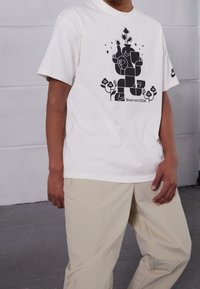 Bílý bavlněný tričko s černým grafickým designem obsahujícím abstraktní tvary a květiny, spárované s béžovými kalhotami. Logo Nike na rukávu.