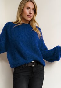 Vrouw met lang blond haar, gekleed in een loszittende, gestructureerde blauwe trui en zwarte spijkerbroek met een zwarte riem, in een neutrale binnenomgeving.