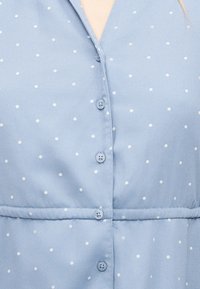 Svetlo modra tkanina z majhnimi belimi pikami, z štirimi prišitimi gumbi in šivom po sredini.