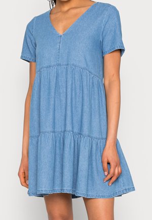 Nainen, joka on pukeutunut lyhythihaiseen, vaaleansiniseen farkkumekkoon, jossa on V-aukko ja kerroksittain laskeutuva helma, seisoo yksinkertaisen vaalean taustan edessä.