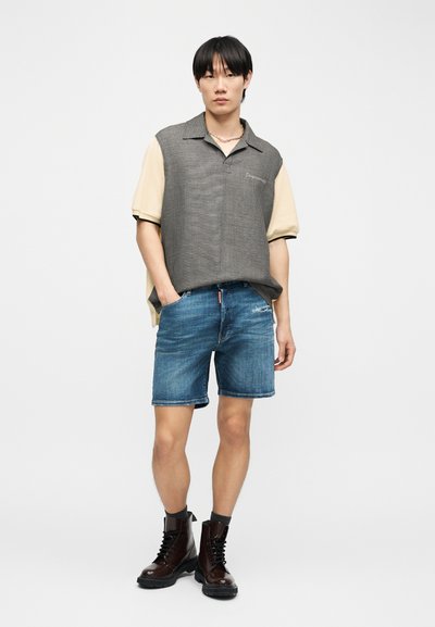 Νέος άνδρας που φοράει μπεζ πουκάμισο με γκρι καρό γιλέκο, μπλε τζιν σορτς, γκρι κάλτσες και σκούρα καφέ μποτάκια με κορδόνια.