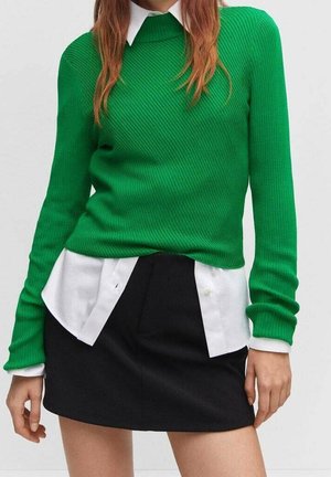 Femme portant un pull vert côtelé à manches longues sur une chemise blanche à col et une mini-jupe noire.