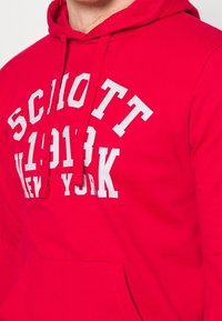 Felpa con cappuccio rossa in morbido tessuto, con una stampa bianca in grassetto con la scritta "SCHOTT 1913 NEW YORK." Include una tasca frontale e un cordoncino.