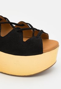 Sandale à plateforme en daim noir avec lacets croisés et semelle beige épaisse et texturée, design à bout ouvert sur fond blanc.