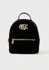 Black velvet backpack with a rounded shape, gold monogram logo on the front, and a zippered front pocket with gold hardware.