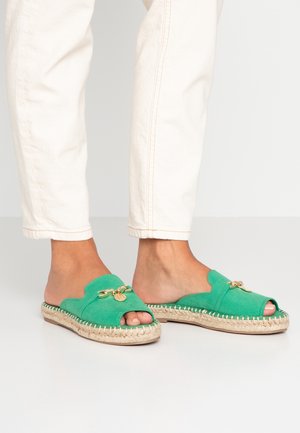 Stopy w zielonych zamszowych sandałach espadrylach typu slip-on z odkrytymi palcami, ozdobionych złotym łańcuszkiem i beżowymi podeszwami z plecionej liny, zestawione z kremowymi spodniami.