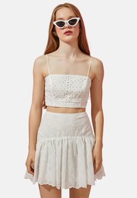Top corto blanco con diseño de ojales, acompañado de una falda de volantes a juego. Presenta tirantes finos y un dobladillo acampanado, con una textura suave.