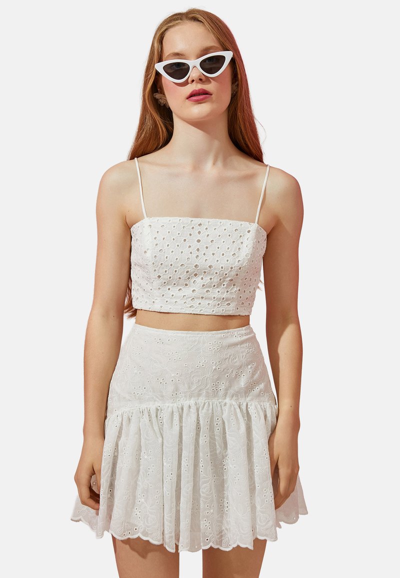 Top corto blanco con diseño de ojales, acompañado de una falda de volantes a juego. Presenta tirantes finos y un dobladillo acampanado, con una textura suave.