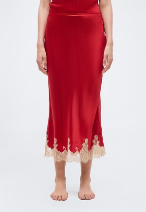Red satin midi skirt with beige lace trim at hem worn by a barefoot person, hands relaxed at sides against a plain background.