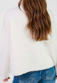 Długie falowane brązowe włosy na białym teksturowanym swetrze w połączeniu z niebieskimi dżinsami, widok z tyłu.