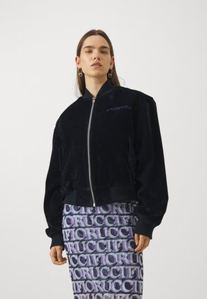 Czarna aksamitna kurtka bomberka z zamkiem błyskawicznym, ściągaczami na rękawach i w pasie, z haftem "Fiorucci" w kolorze purpurowym na klatce piersiowej.