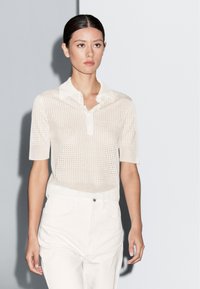 Witte gebreide poloshirt met korte mouwen, met een gestructureerd patroon. Model draagt lichtgekleurde broek, tegen een neutrale achtergrond.