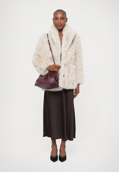 Manteau en fausse fourrure beige clair avec un large col, associé à une robe midi marron foncé. Accessoirisé avec un sac bordeaux et des escarpins noirs.
