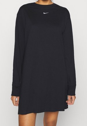 Frau trägt ein langärmliges, schwarzes Nike-Kleid mit kleinem weißem Swoosh-Logo auf der Brust und steht vor einem einfarbigen Hintergrund.