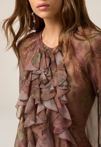 Durchsichtige Blumenbluse in sanftem Pink und Grün, mit geschichteten Rüschen entlang der Vorderseite, runder Ausschnitt und zarten Stickdetails.