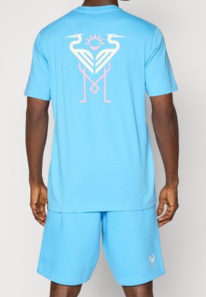 Homme portant un short bleu vif et un T-shirt avec un dessin abstrait d'oiseau rose et blanc et de soleil au dos et sur le short.