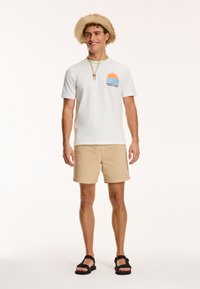 Tricou alb din bumbac cu imprimeu grafic de soare și val. Șorturi bej și sandale negre. Pălărie din paie cu boruri curbe și curea.