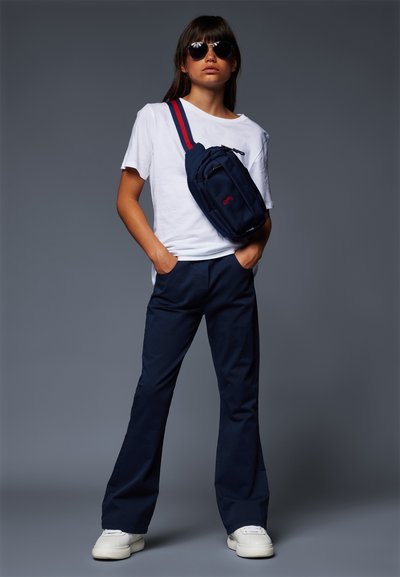 Saco crossbody azul-marinho com alça vermelha e azul, camiseta branca e calças azul-marinho. Modelo usando óculos de sol, de pé de forma confiante com as mãos nos bolsos.