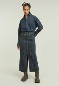 Camisa de denim con cierre de botones, mangas largas y un lavado oscuro. La falda de cintura alta presenta una abertura frontal y detalles de costura en contraste.