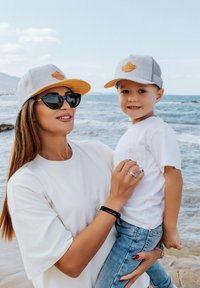 Grau gestreifte Mütze mit orangefarbenen Akzenten und Patch, kombiniert mit einem weißen T-Shirt und Jeansshorts, vor einem Strandhintergrund mit Meereswellen.